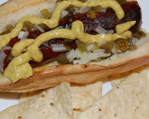 Prepared hot dog with mustard, diced onion, jalapeño and relish on a white plate.