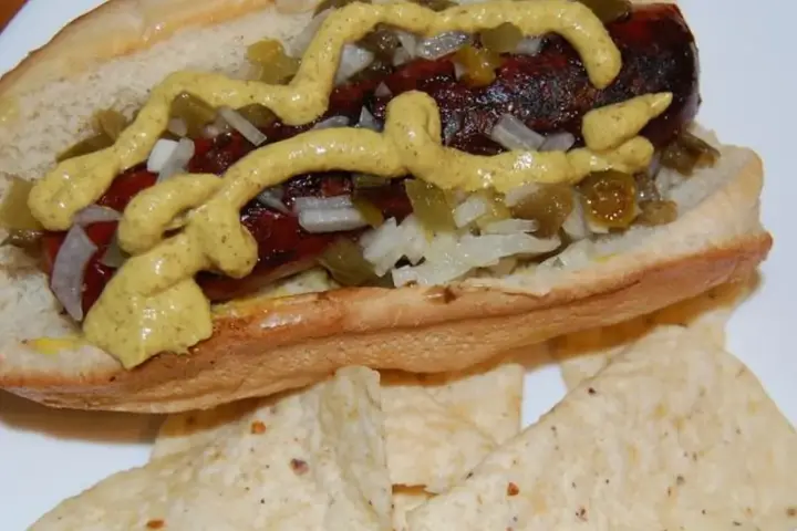 Prepared hot dog with mustard, diced onion, jalapeño and relish on a white plate.