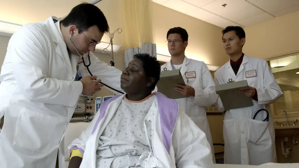 Photograph of a physician in clinical attire using a stethoscope to examine a patient in an outpatient consultation room while colleagues review notes.