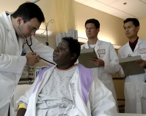Photograph of a physician in clinical attire using a stethoscope to examine a patient in an outpatient consultation room while colleagues review notes.