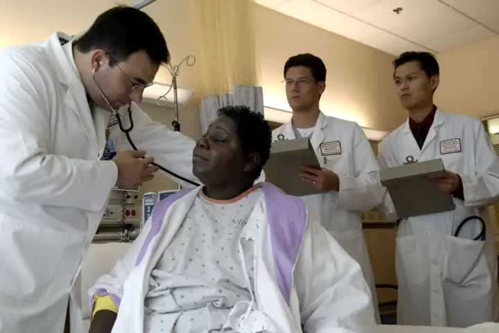 Photograph of a physician in clinical attire using a stethoscope to examine a patient in an outpatient consultation room while colleagues review notes.