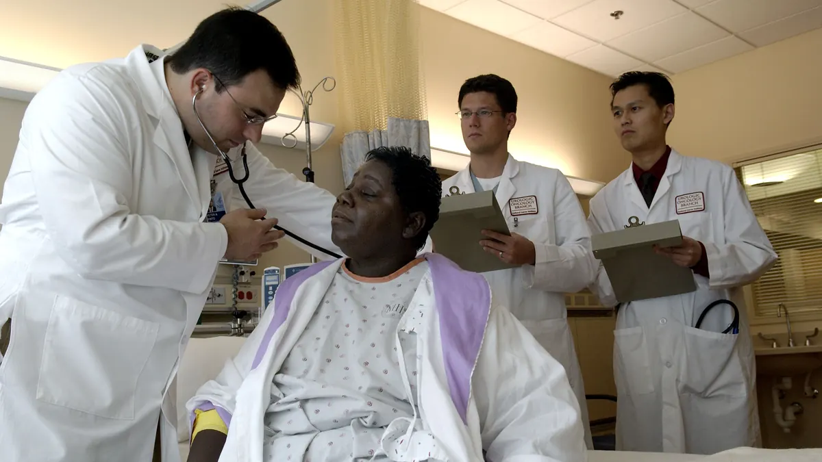 Photograph of a physician in clinical attire using a stethoscope to examine a patient in an outpatient consultation room while colleagues review notes.
