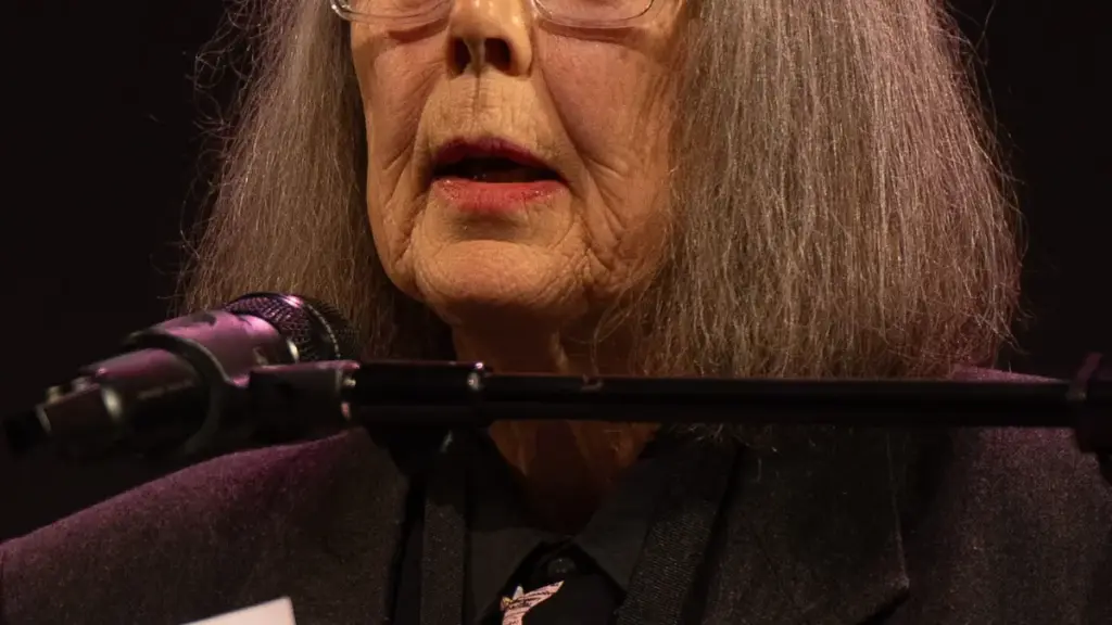 Portrait photograph of Canadian author and translator Anne Carson wearing glasses and addressing a seated audience at a literary event.