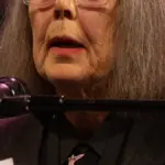 Portrait photograph of Canadian author and translator Anne Carson wearing glasses and addressing a seated audience at a literary event.