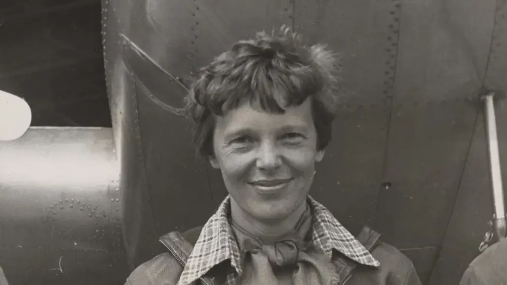Historical portrait photograph of aviator Amelia Earhart standing beneath the engine and fuselage of her twin-engine Lockheed Model 10-E Electra aircraft.