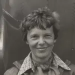 Historical portrait photograph of aviator Amelia Earhart standing beneath the engine and fuselage of her twin-engine Lockheed Model 10-E Electra aircraft.