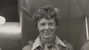 Historical portrait photograph of aviator Amelia Earhart standing beneath the engine and fuselage of her twin-engine Lockheed Model 10-E Electra aircraft.