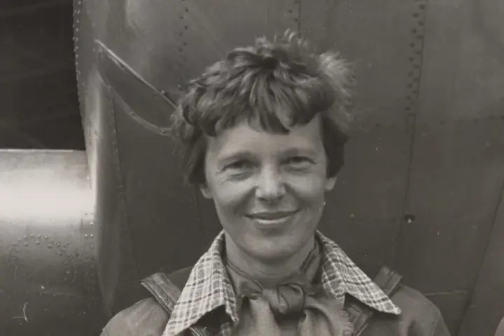 Historical portrait photograph of aviator Amelia Earhart standing beneath the engine and fuselage of her twin-engine Lockheed Model 10-E Electra aircraft.
