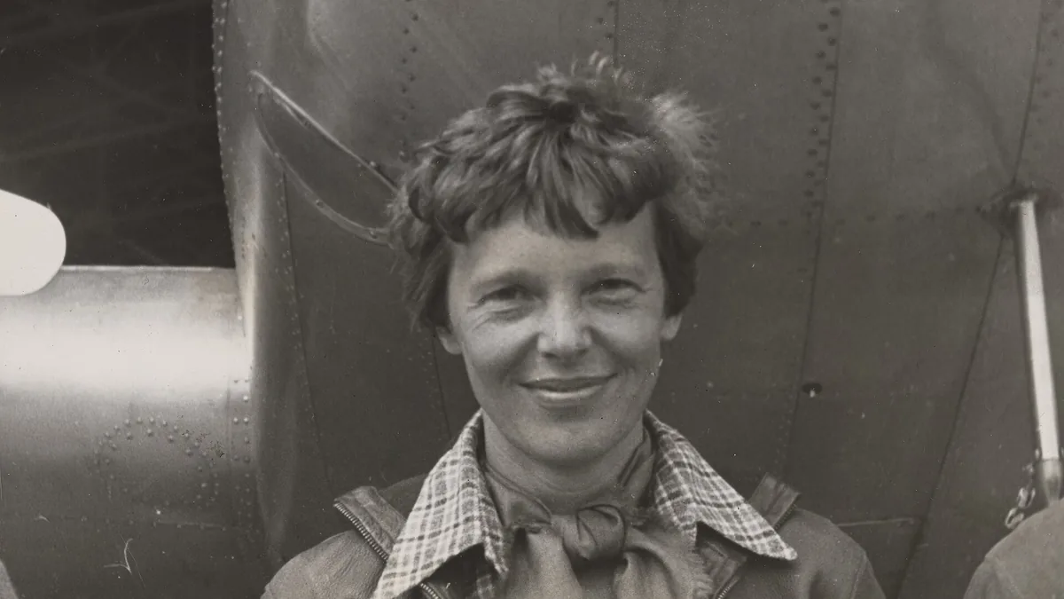 Historical portrait photograph of aviator Amelia Earhart standing beneath the engine and fuselage of her twin-engine Lockheed Model 10-E Electra aircraft.