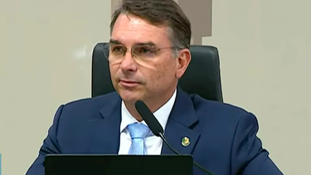 Brazilian Senator Flávio Bolsonaro wearing glasses, a dark navy suit, and a light blue tie seated behind a microphone at the Federal Senate in Brasília.