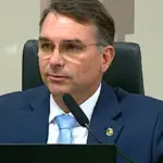 Brazilian Senator Flávio Bolsonaro wearing glasses, a dark navy suit, and a light blue tie seated behind a microphone at the Federal Senate in Brasília.