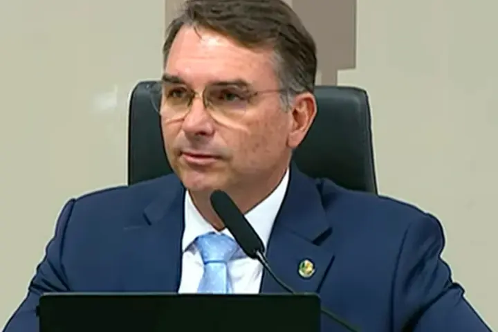 Brazilian Senator Flávio Bolsonaro wearing glasses, a dark navy suit, and a light blue tie seated behind a microphone at the Federal Senate in Brasília.