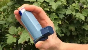 Close-up photograph of a hand holding a blue plastic metered-dose asthma inhaler outdoors in front of green leaves.