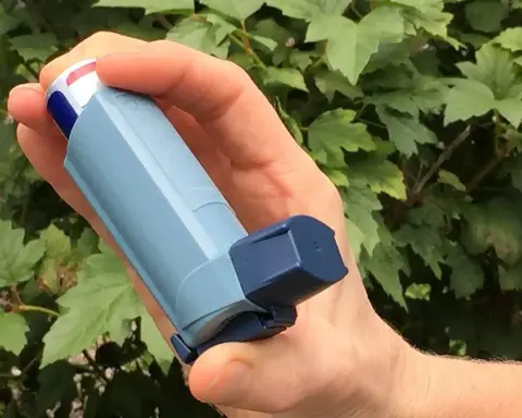 Close-up photograph of a hand holding a blue plastic metered-dose asthma inhaler outdoors in front of green leaves.
