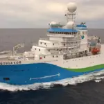 Port-side aerial view of the blue, white, and green CSIRO research vessel RV Investigator steaming through deep ocean waters off Tasmania.