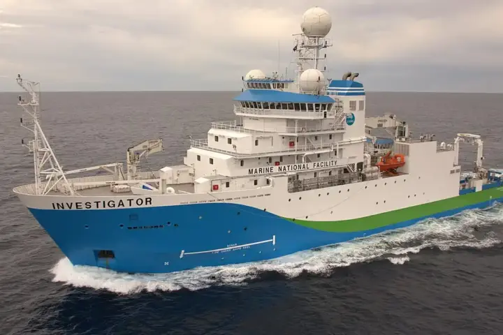 Port-side aerial view of the blue, white, and green CSIRO research vessel RV Investigator steaming through deep ocean waters off Tasmania.