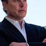 Portrait of Elon Musk wearing a black suit jacket and white open-collar shirt with his arms crossed outdoors in front of a mountain ridge.