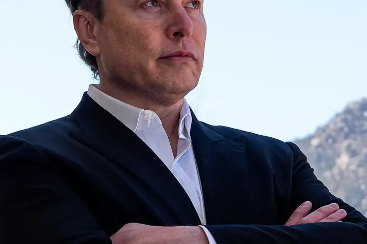 Portrait of Elon Musk wearing a black suit jacket and white open-collar shirt with his arms crossed outdoors in front of a mountain ridge.