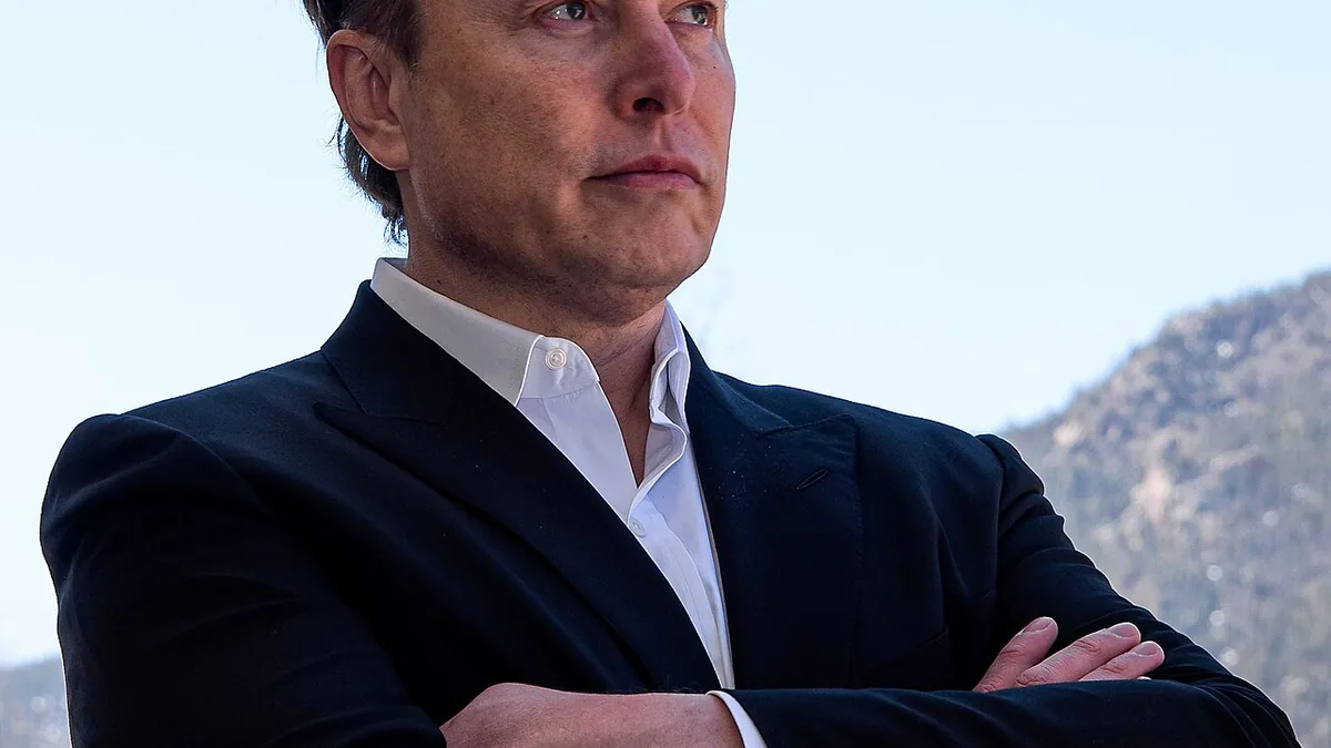 Portrait of Elon Musk wearing a black suit jacket and white open-collar shirt with his arms crossed outdoors in front of a mountain ridge.