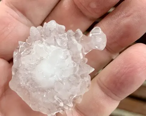 Close-up photograph of a person holding a large, spiky, translucent and white hailstone over a wooden deck in South-East Queensland.