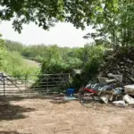 Pile of illegally dumped commercial and construction rubbish next to a metal farm gate on a tree-lined dirt track in a rural area.