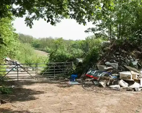 Pile of illegally dumped commercial and construction rubbish next to a metal farm gate on a tree-lined dirt track in a rural area.