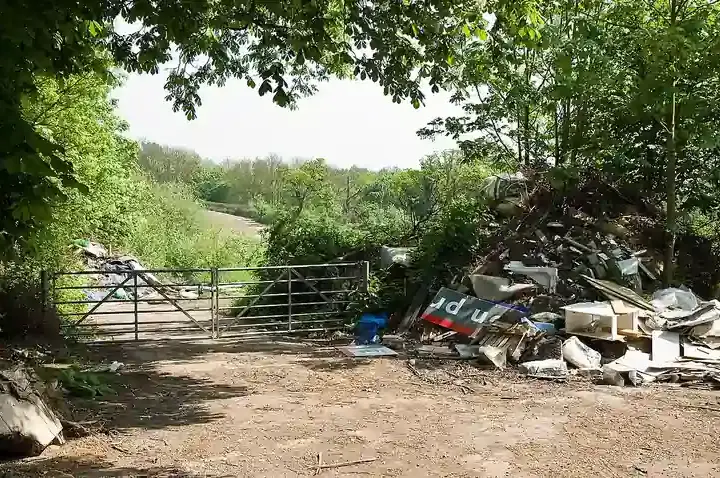 Pile of illegally dumped commercial and construction rubbish next to a metal farm gate on a tree-lined dirt track in a rural area.