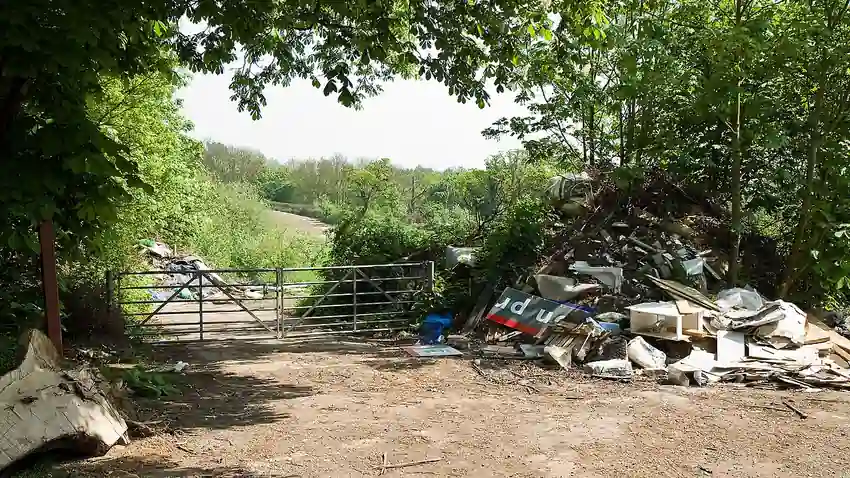 Pile of illegally dumped commercial and construction rubbish next to a metal farm gate on a tree-lined dirt track in a rural area.