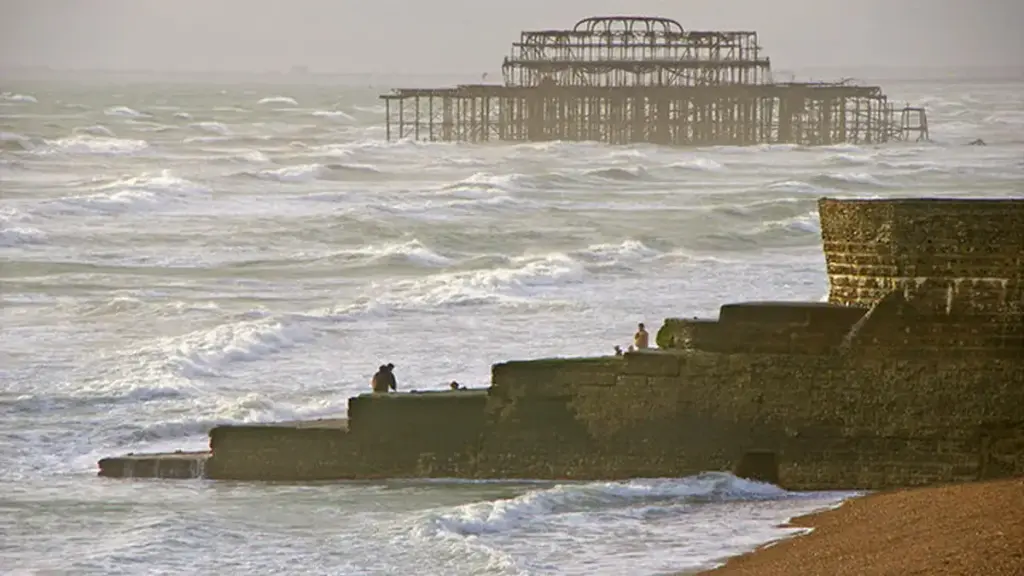 Rough waves breaking over a concrete beach groyne in Brighton, England, with the skeletal ruins of the West Pier visible offshore.