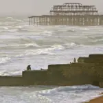 Rough waves breaking over a concrete beach groyne in Brighton, England, with the skeletal ruins of the West Pier visible offshore.