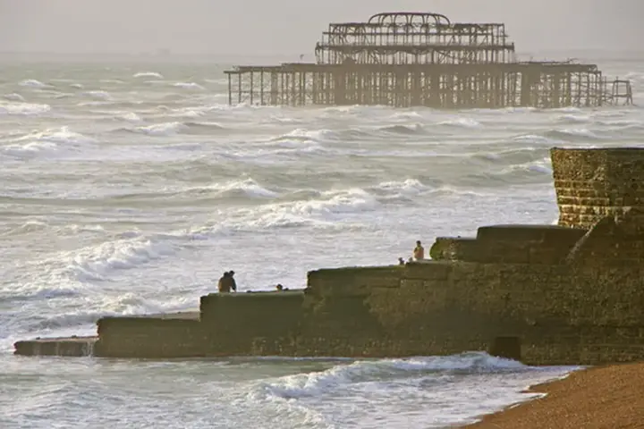 Rough waves breaking over a concrete beach groyne in Brighton, England, with the skeletal ruins of the West Pier visible offshore.