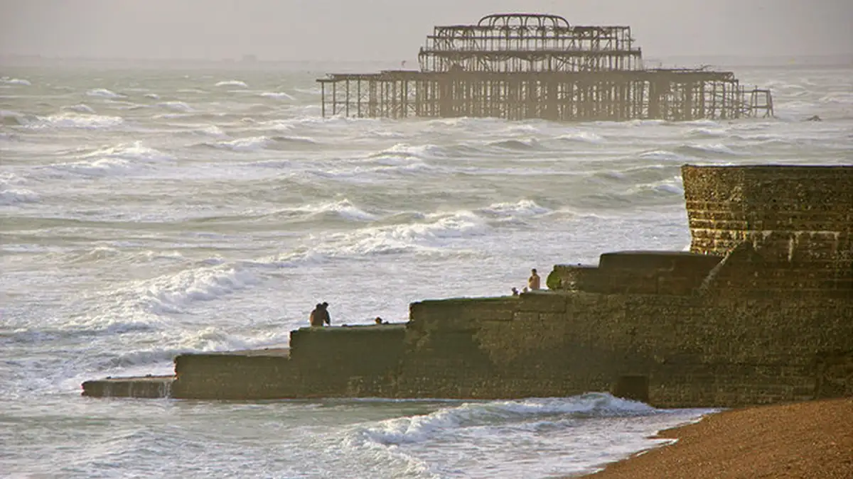 Rough waves breaking over a concrete beach groyne in Brighton, England, with the skeletal ruins of the West Pier visible offshore.