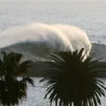 Powerful ocean wave cresting and breaking heavily against the sandy shoreline near coastal structures in Newport Beach, California.
