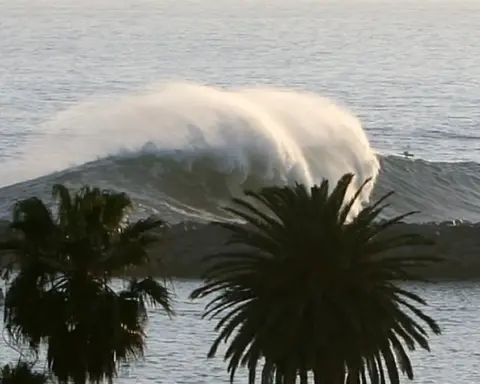 Powerful ocean wave cresting and breaking heavily against the sandy shoreline near coastal structures in Newport Beach, California.