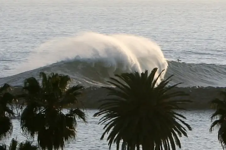 Powerful ocean wave cresting and breaking heavily against the sandy shoreline near coastal structures in Newport Beach, California.