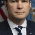 Formal photographic portrait of US Secretary of Defense Pete Hegseth standing before the United States and Department of Defense flags.