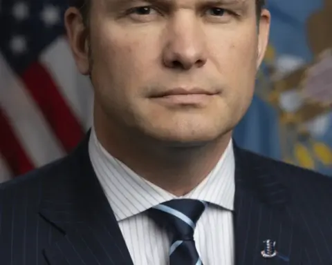 Formal photographic portrait of US Secretary of Defense Pete Hegseth standing before the United States and Department of Defense flags.