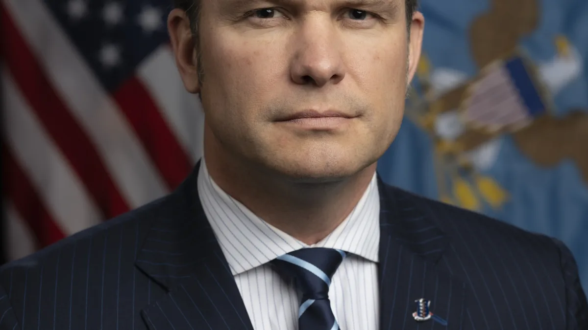Formal photographic portrait of US Secretary of Defense Pete Hegseth standing before the United States and Department of Defense flags.