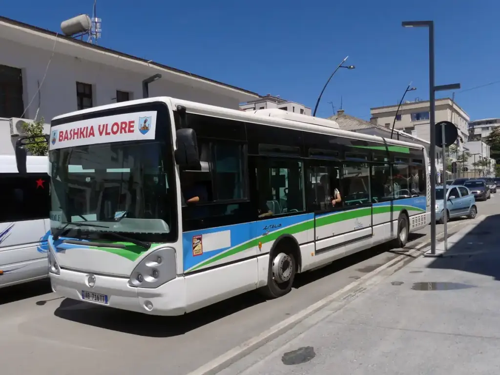 White Citelis city bus in Vlora, Albania, on a sunny urban street with parked cars, buildings, and clear blue sky.