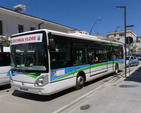 White Citelis city bus in Vlora, Albania, on a sunny urban street with parked cars, buildings, and clear blue sky.