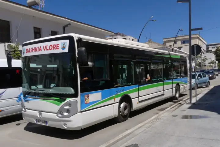 White Citelis city bus in Vlora, Albania, on a sunny urban street with parked cars, buildings, and clear blue sky.