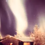 Green and purple aurora borealis forming luminous curtains in the night sky above a snow-covered cabin in Alaska