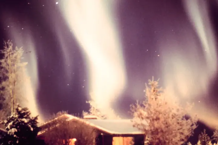 Green and purple aurora borealis forming luminous curtains in the night sky above a snow-covered cabin in Alaska