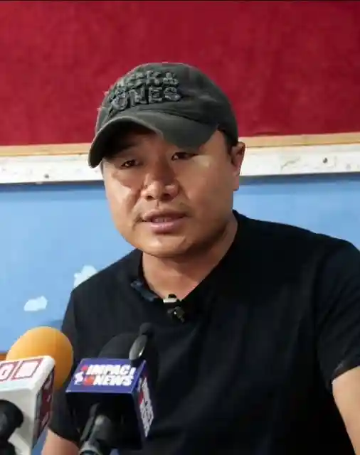 Man speaking at press conference with Impact News microphone flags