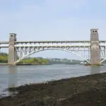 Britannia Bridge and Menai Strait under a cloudy sky, with green hills in the background, North Wales.