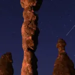 Bright meteor streaks across a dark night sky over a rocky desert landscape with star trails visible