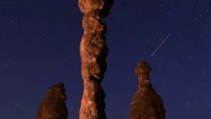 Bright meteor streaks across a dark night sky over a rocky desert landscape with star trails visible