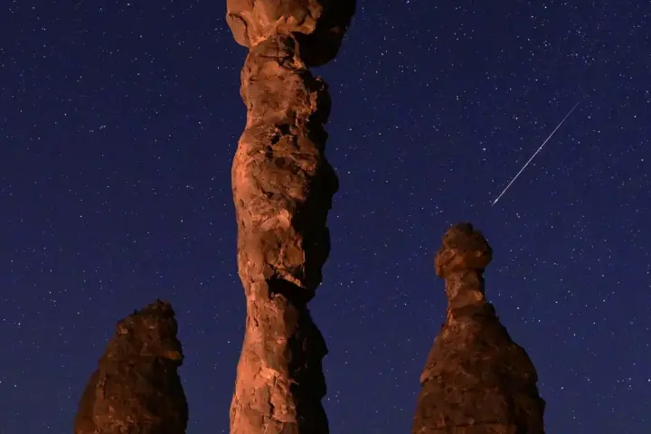 Bright meteor streaks across a dark night sky over a rocky desert landscape with star trails visible