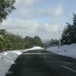 A winding mountain road cutting through snow-covered forest and rocky terrain under a pale sky