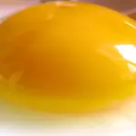 A fresh cracked raw egg resting on a clean surface with a prominent intact golden yolk and clear egg white.